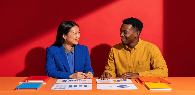 Two colleagues smile at each other at a table with charts, against a bold red background. Woman in blue blazer, man in yellow shirt.